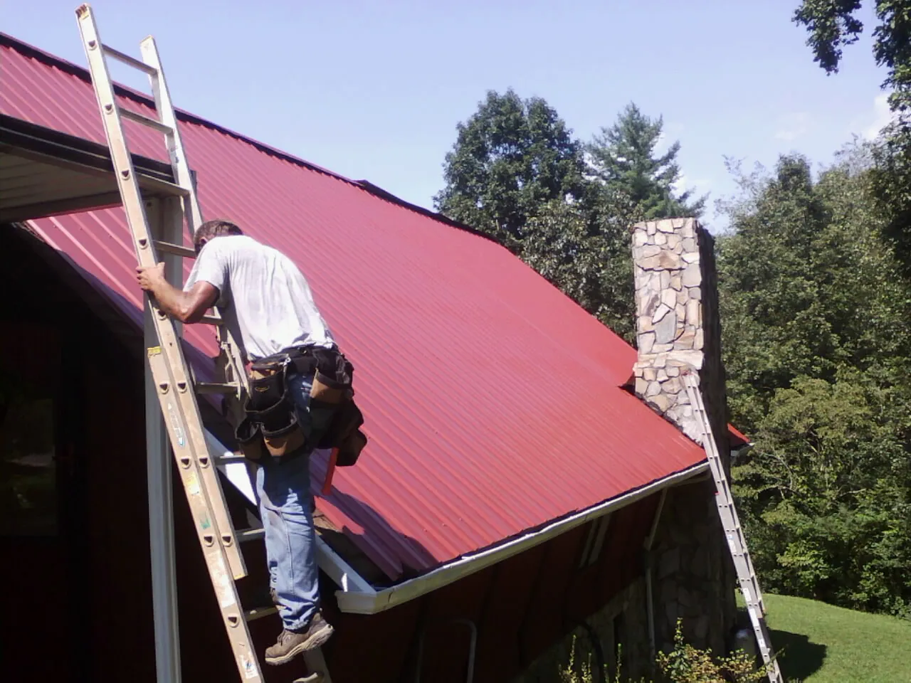 Roofer on a ladder beside a red metal roof and stone chimney — Soffit & Fascia Repair by Roofing Brothers in Boykins